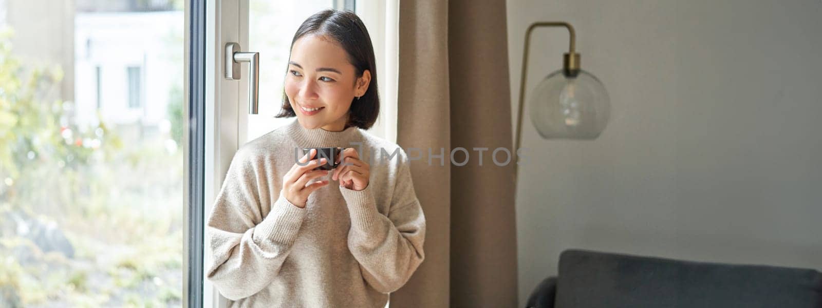 Portrait of beautiful asian woman sitting at home with cup of coffee, enjoying her espresso while looking outside window at passerby, smiling happily.