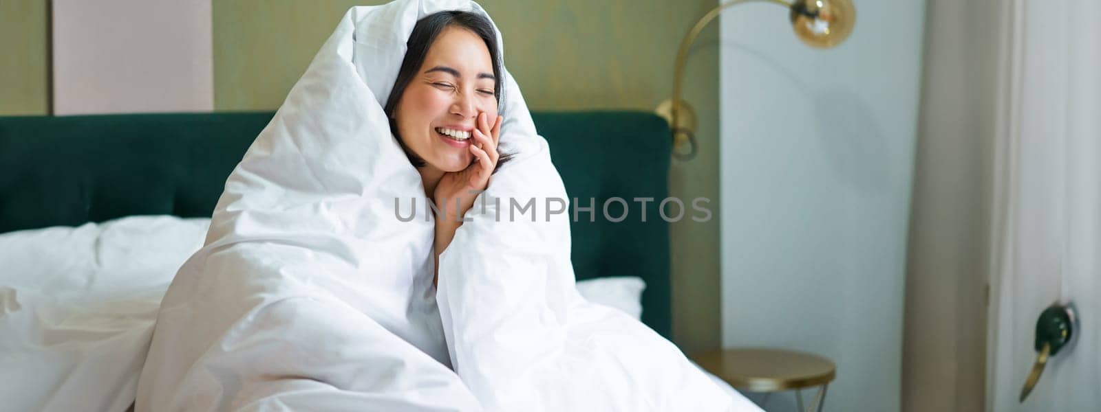 Beautiful asian woman sitting on bed, covered in blankets and duvet, laughing and smiling, enjoying weekend in bedroom.