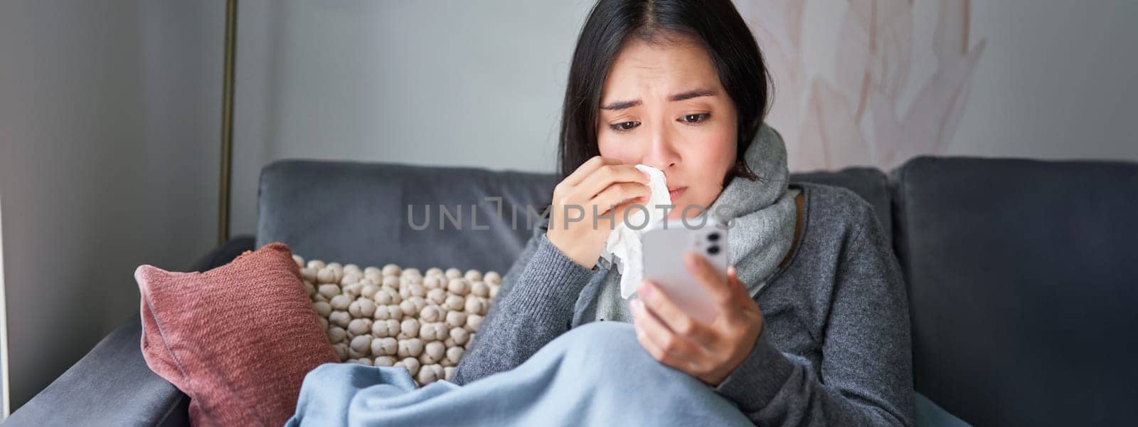 Portrait of sick woman contacting her GP via mobile phone while catching cold, sitting with flu at home, using smartphone by Benzoix