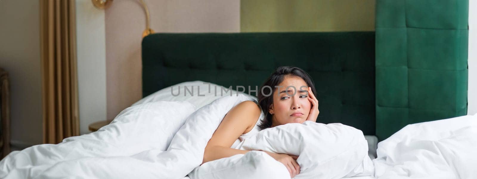 Sad and distressed young korean woman lying in her bed with pillow and warm duvet, feels uneasy, frowning and grimacing by Benzoix