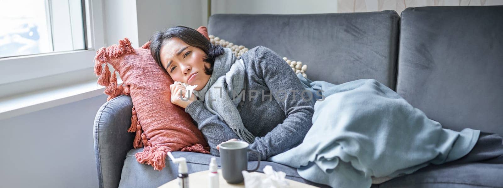 Sick woman lying on sofa at home, catching cold. Young girl freezing from heating problem in her apartment, concept of high cost of living.