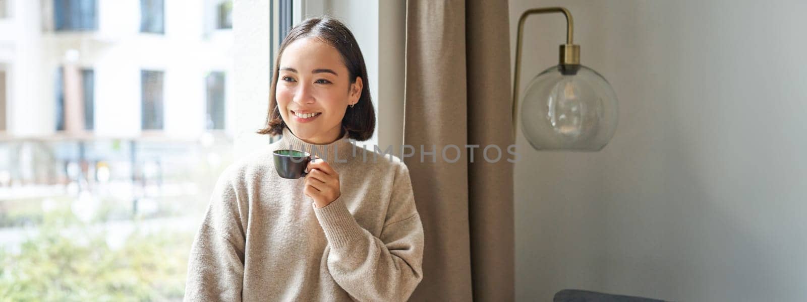 Portrait of beautiful asian woman sitting at home with cup of coffee, enjoying her espresso while looking outside window at passerby, smiling happily by Benzoix