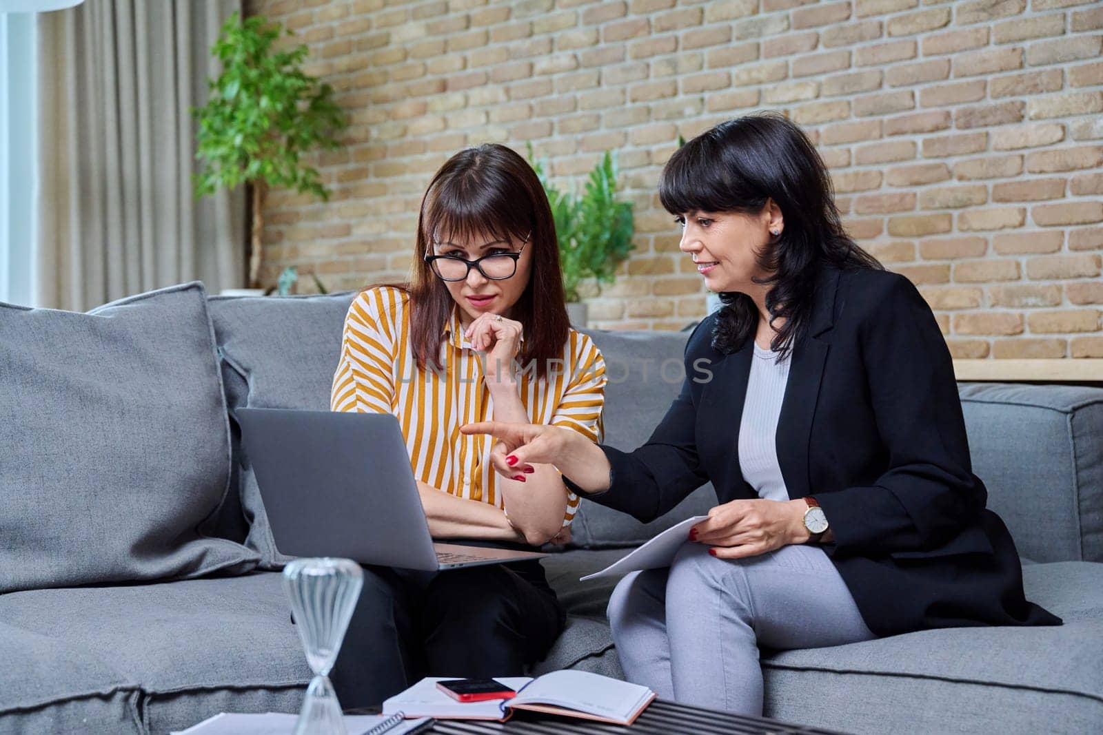 Two mature business women having conversation sitting on couch in office, discussing work, commercial projects. Communicating female colleague, 40s 50s age. Law finance mentoring consulting teamwork