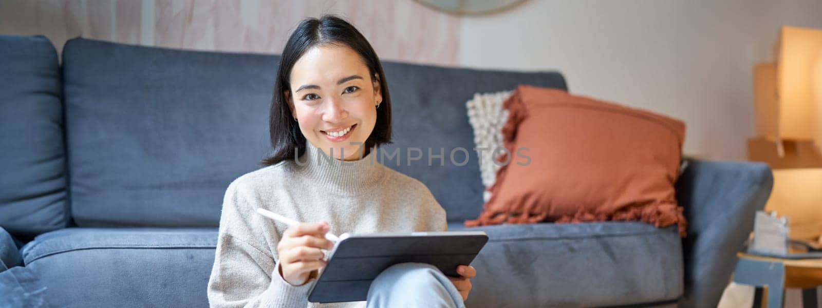 Portrait of asian woman with tablet, drawing, working on design project, holding pen, sitting in her living room, freelancing.