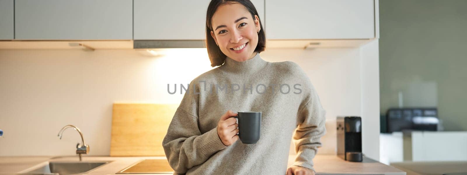 Portrait of smiling asian woman standing on her kitchen, drinking coffee and looking at camera, concept of cozy and comfortable home.