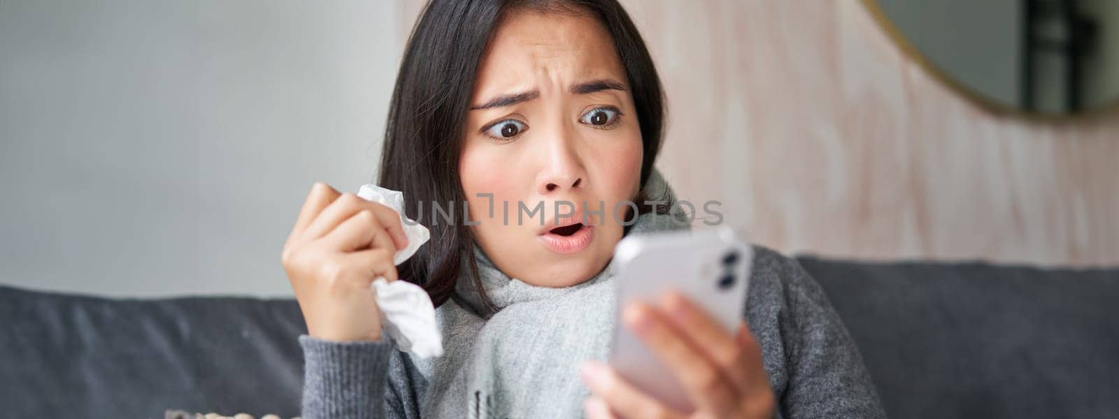 Portrait of ill woman looking at smartphone, looking concerned and shocked at mobile phone, reading bad news while sitting on sick leave at home by Benzoix