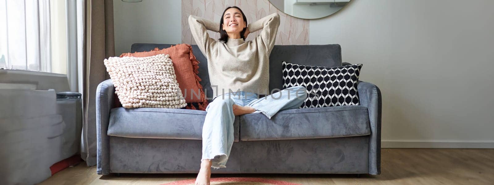 Portrait of carefree asian woman enjoying day-off, sitting on sofa and smiling pleased, relaxing in house on weekends.