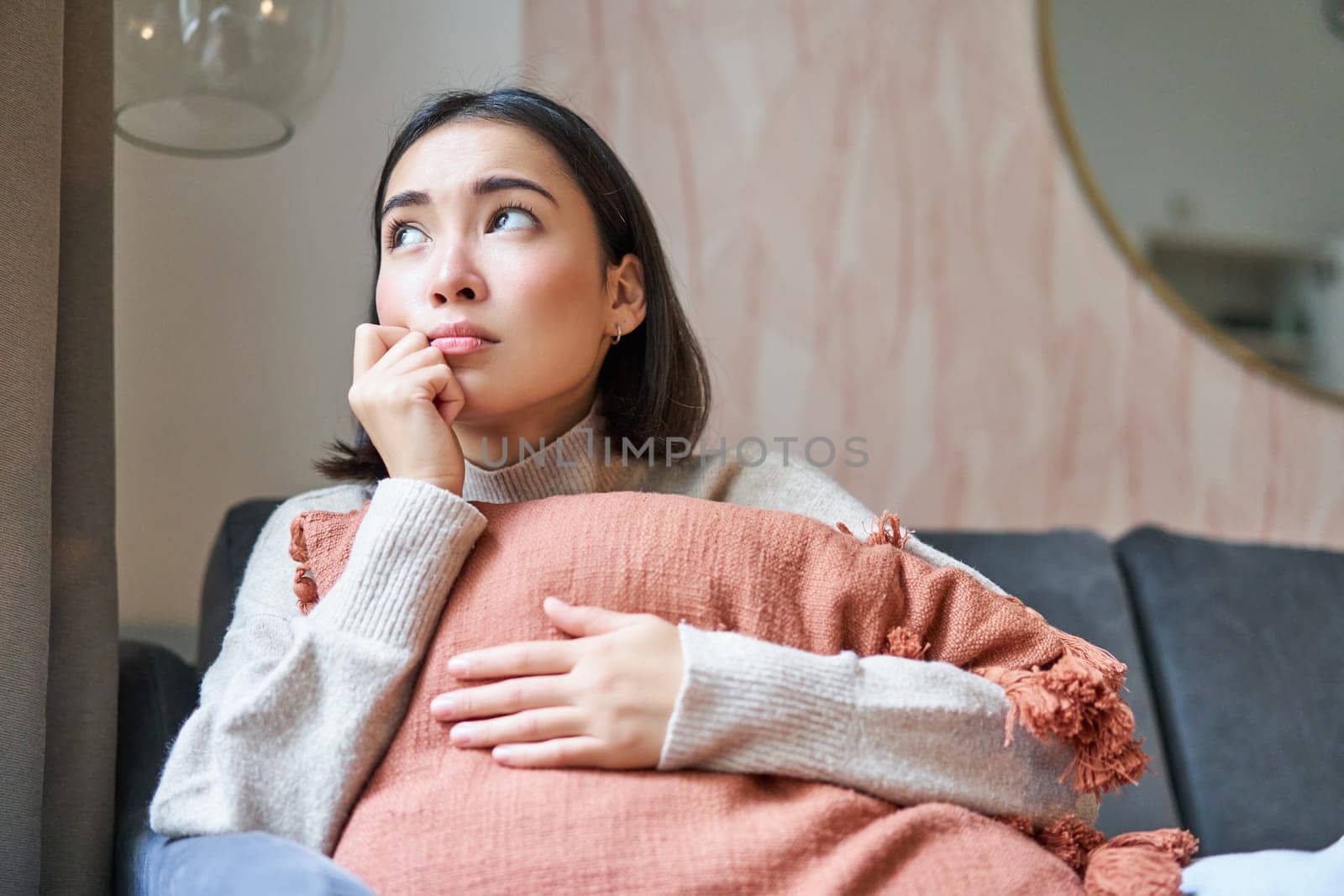 Women and wellbeing concept. Portrait of sad and gloomy asian woman thinking of something unhappy, hugging pillow on sofa and expressing distress by Benzoix