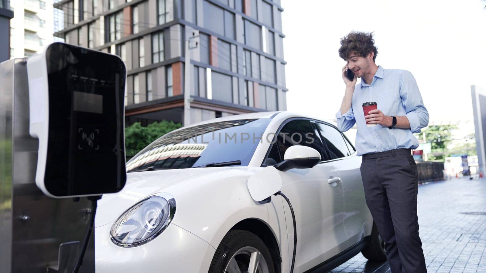 Suit-clad businessman with progressive ambition leaning on his electric vehicle while standing on a charging station with a power cable plug and a renewable energy-powered electric vehicle.