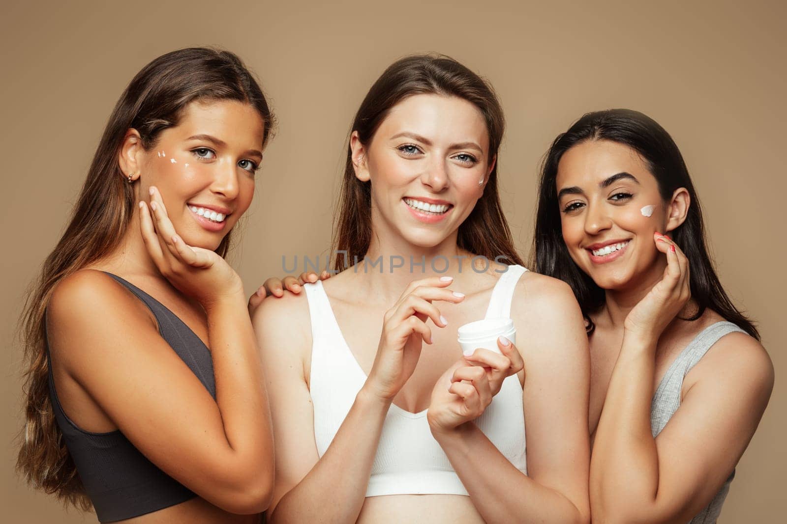 Smiling millennial diverse women in underwear enjoy beauty care, apply cream to face, isolated on beige background. Dermatology, self love, treatments, wellness, skin care, cosmetics, moisturizing