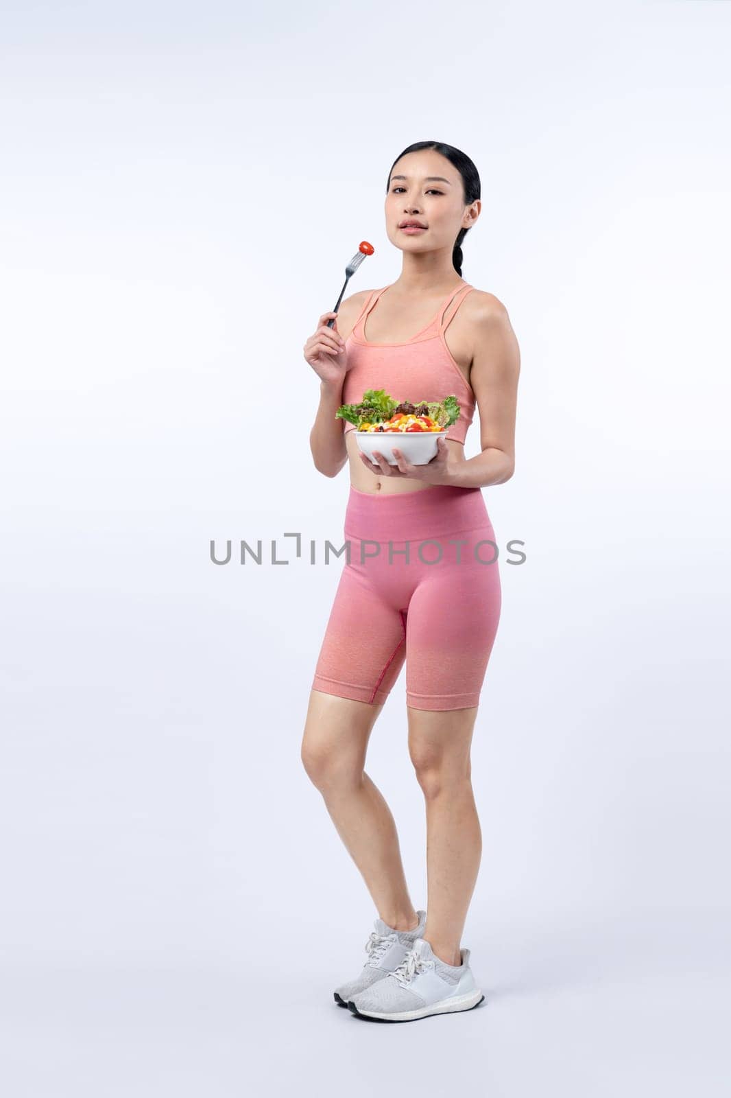 Young sporty Asian woman in sportswear holding salad bowl fill with vibrant of fruit and vegetable. Natural youthful and fit body lifestyle with balance nutrition on isolated background. Vigorous