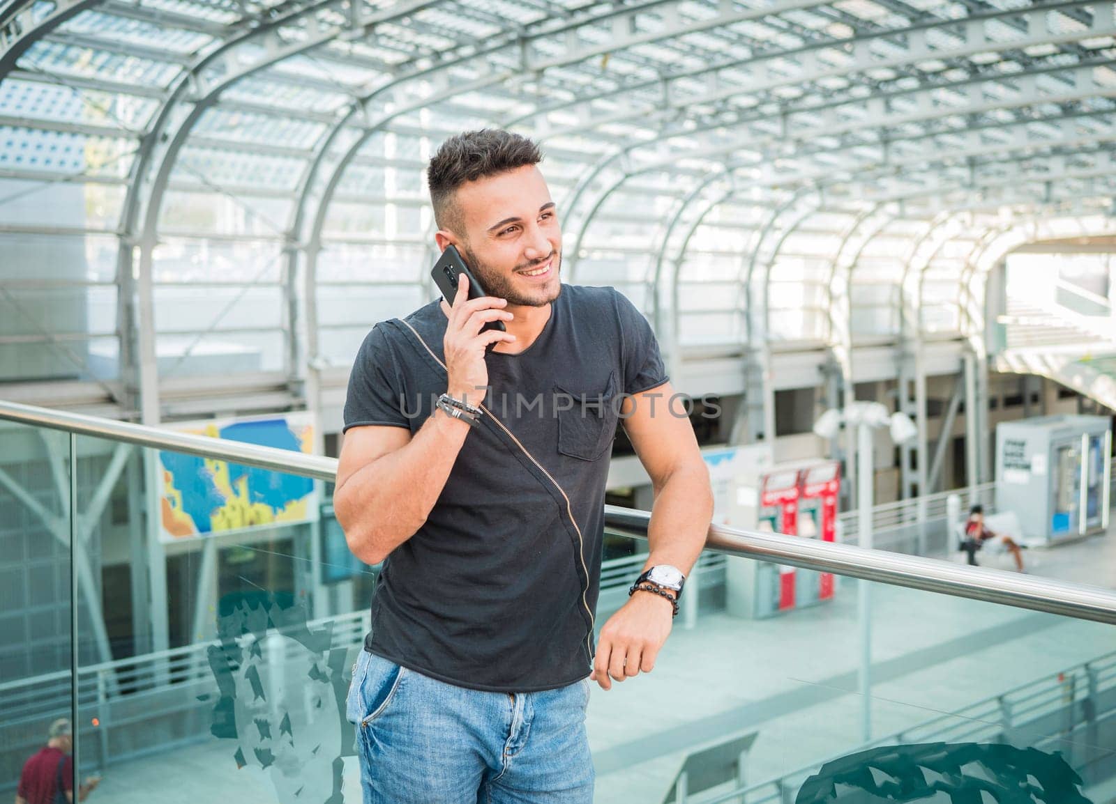 A man standing on a balcony talking on a cell phone. Photo of a young and handsome man talking on a cell phone while standing on a balcony in a modern train station