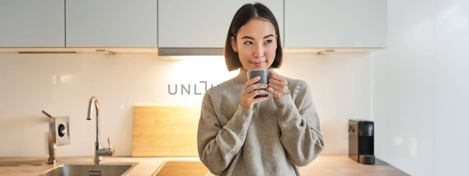 Portrait of smiling asian woman standing on her kitchen, drinking coffee and looking at camera, concept of cozy and comfortable home by Benzoix
