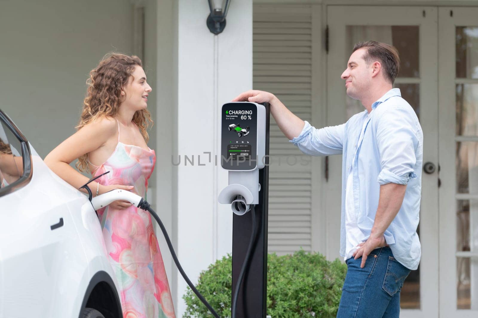 Happy and lovely couple with eco-friendly conscious recharging electric vehicle from EV home charging station. EV car technology utilized for residential home to future sustainability. Synchronos