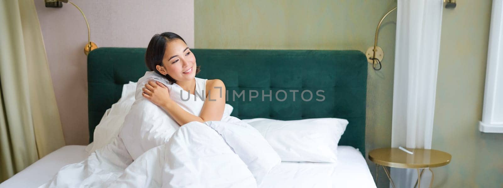 Beautiful sleepy asian girl wakes up in her bed with cozy white blankets, looking outside window with pleased smile. Lifestyle concept.