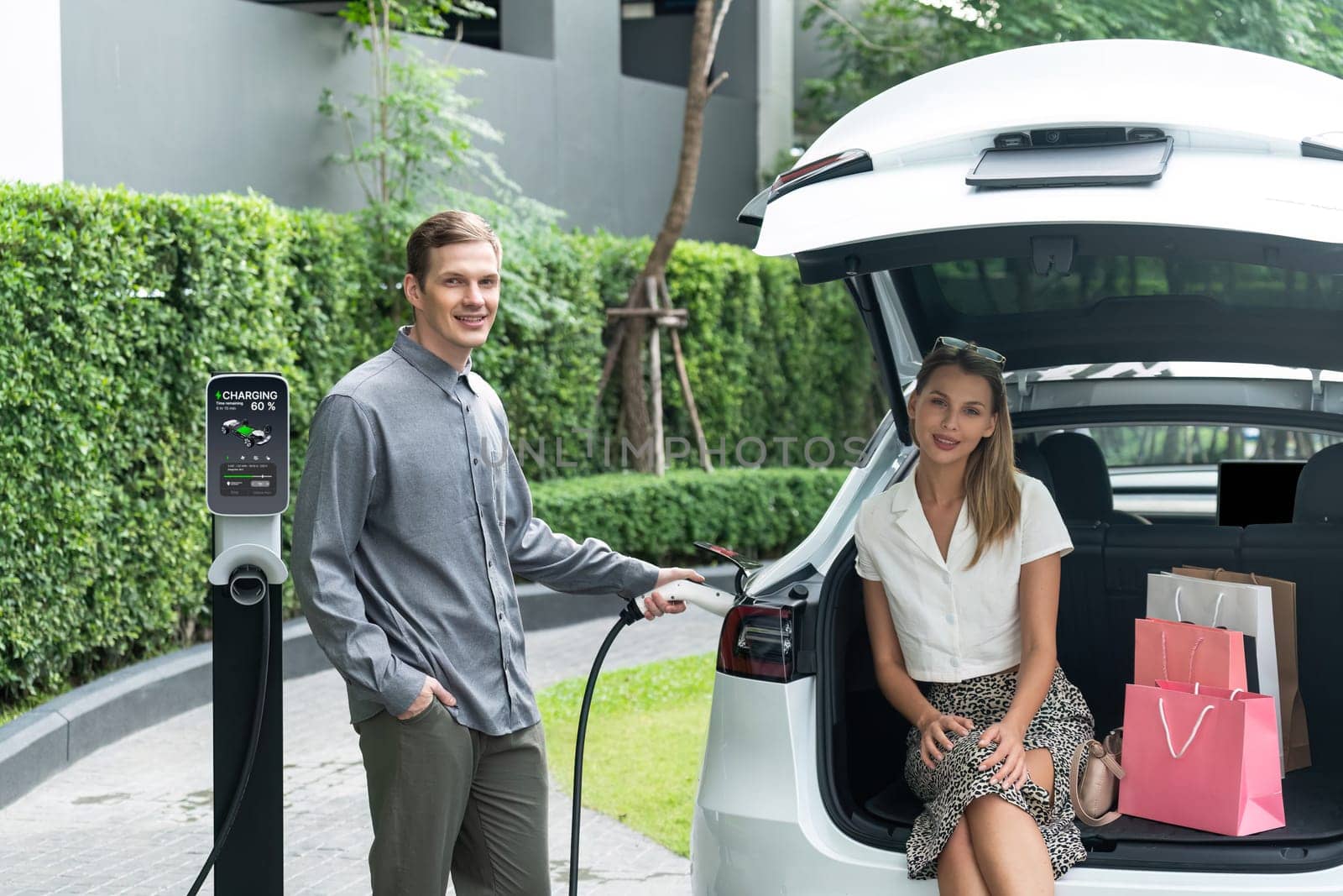 Young couple travel with EV electric car charging in green sustainable city outdoor garden in summer shows urban sustainability lifestyle by green clean rechargeable energy of electric vehicle innards