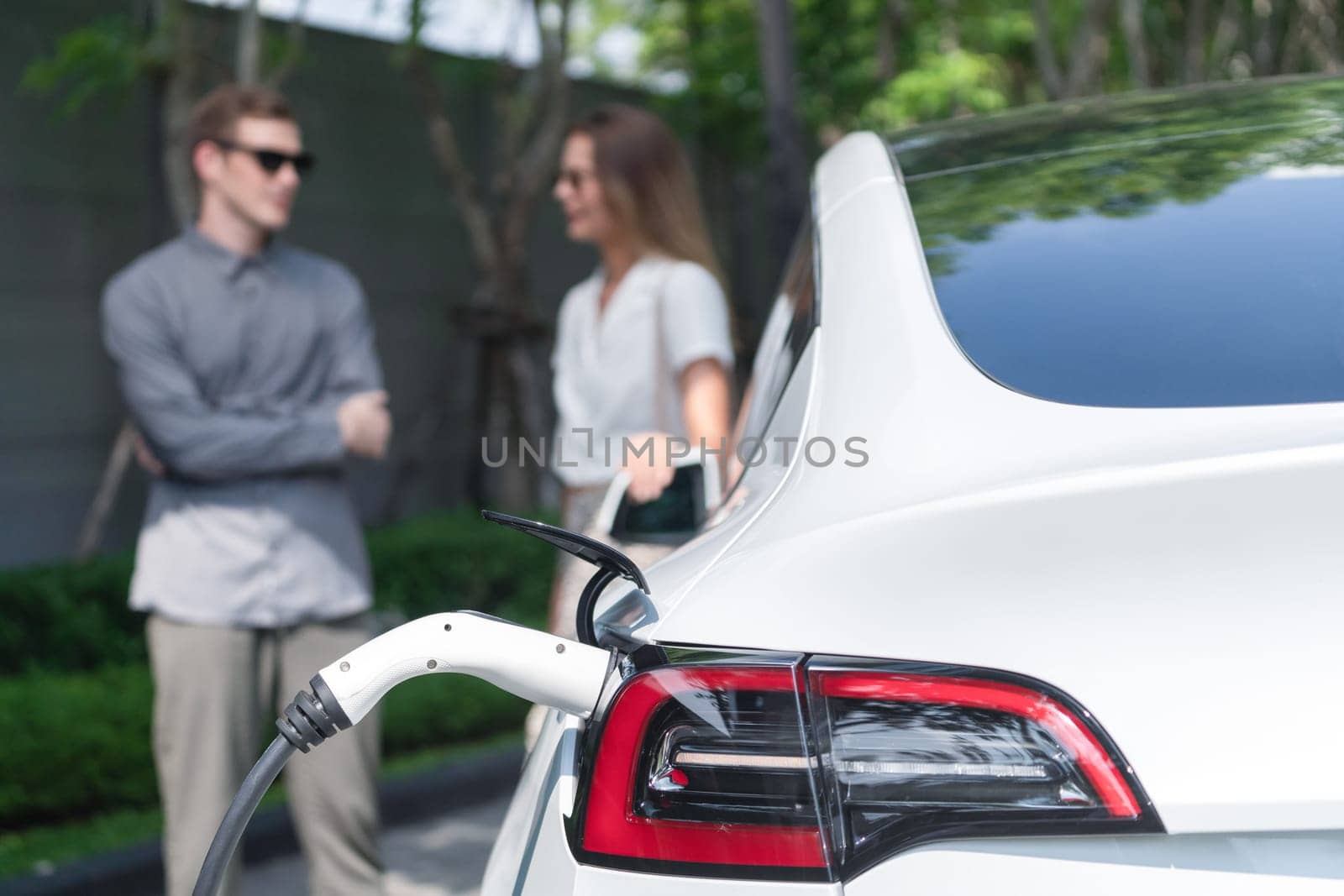 Young couple travel with EV electric car charging in green sustainable city outdoor garden in summer shows urban sustainability lifestyle by green clean rechargeable energy of electric vehicle innards