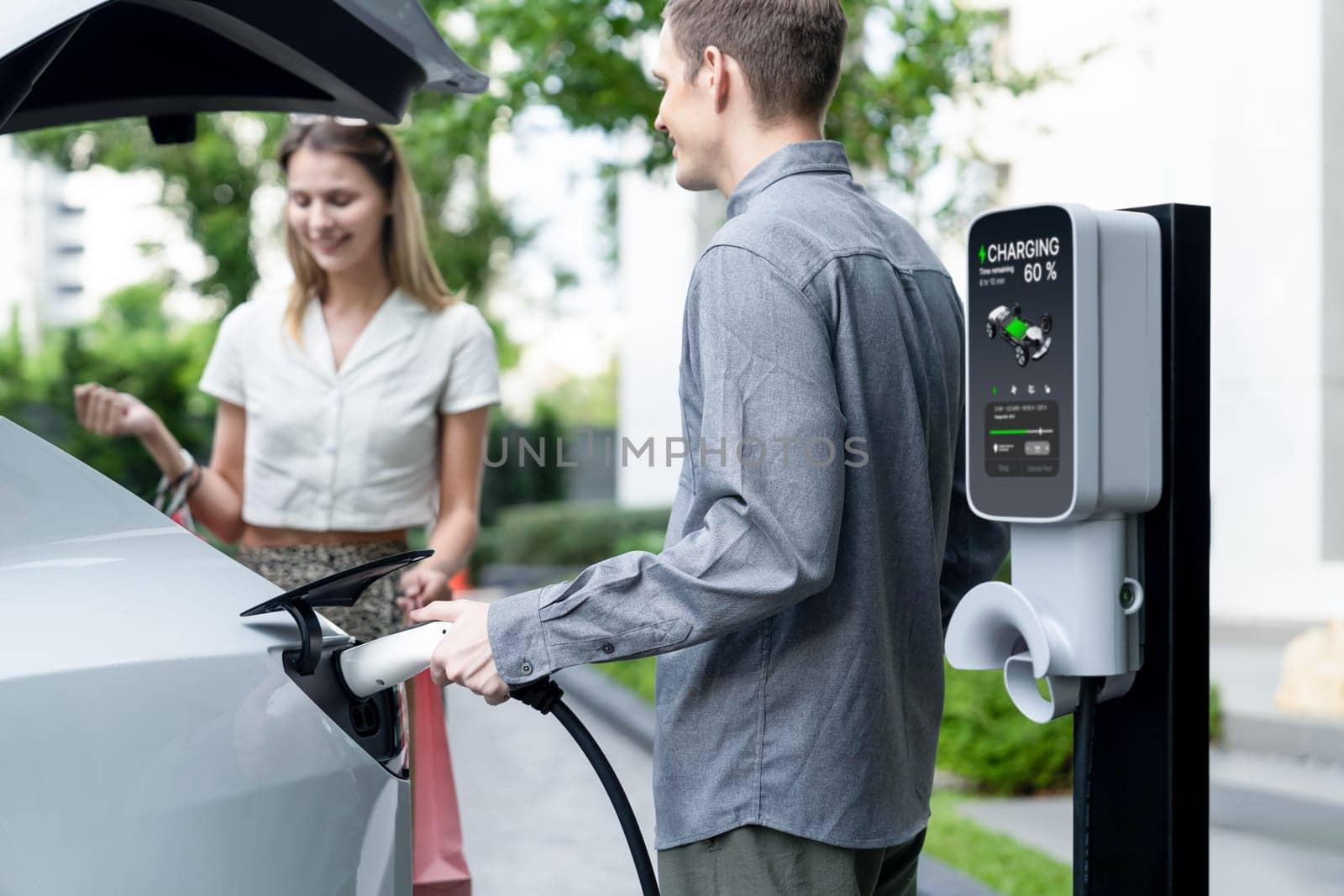 Young couple travel with EV electric car charging in green sustainable city outdoor garden in summer shows urban sustainability lifestyle by green clean rechargeable energy of electric vehicle innards