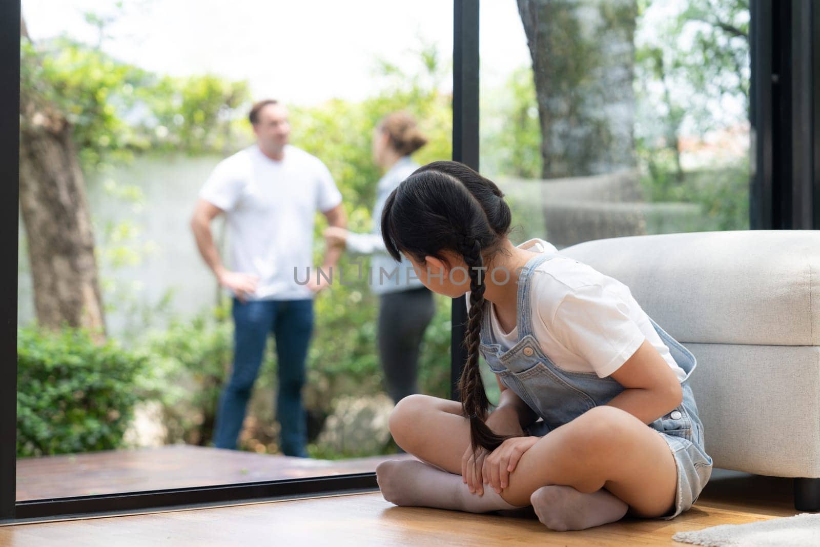 Stressed and unhappy young girl huddle in corner crying and sad while her parent arguing in background. Domestic violence at home and traumatic childhood develop to depression. Synchronos