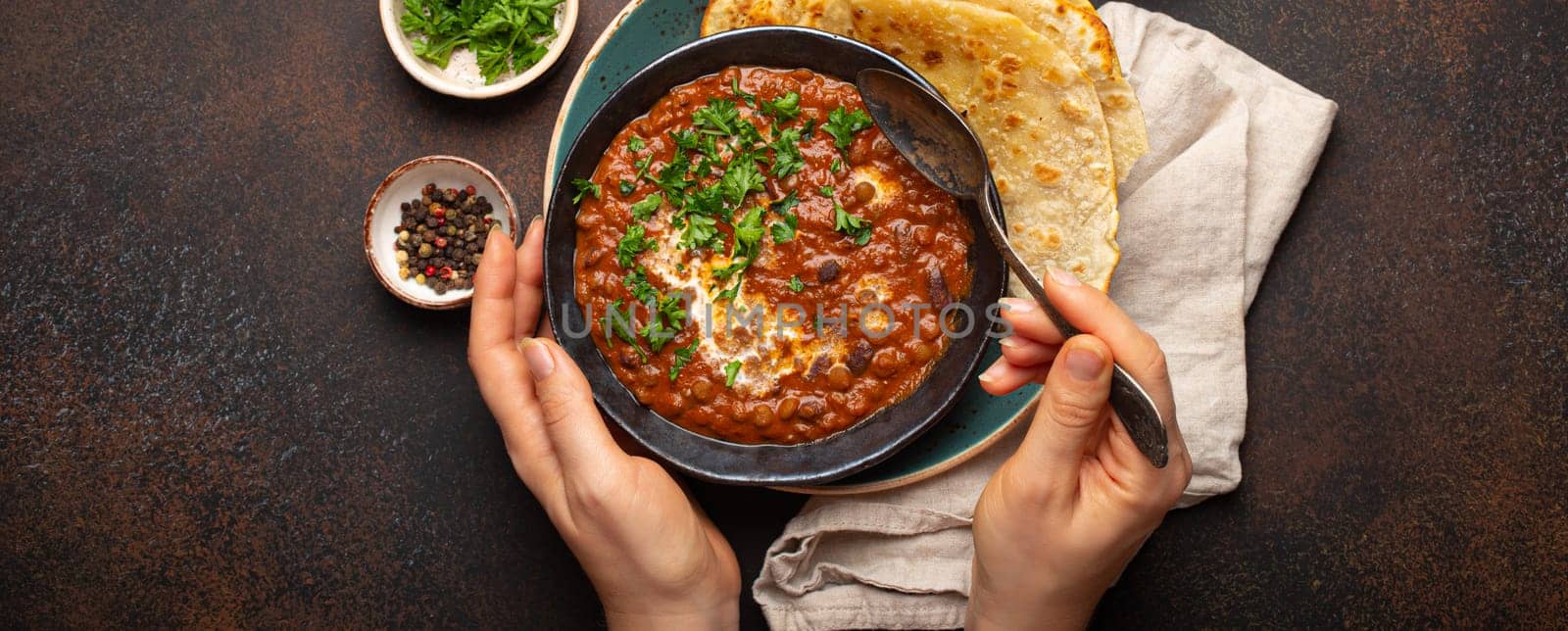 Female hands holding a bowl and eating traditional Indian Punjabi dish Dal makhani with lentils and beans served with naan flat bread, fresh cilantro on brown concrete rustic table top view by its_al_dente