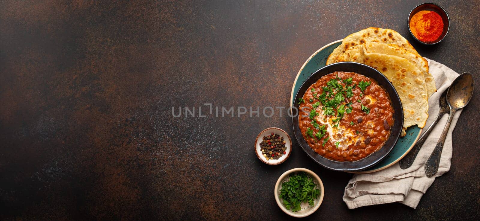 Traditional Indian Punjabi dish Dal makhani with lentils and beans in black bowl served with naan flat bread, fresh cilantro and two spoons on brown concrete rustic table top view. Space for text by its_al_dente