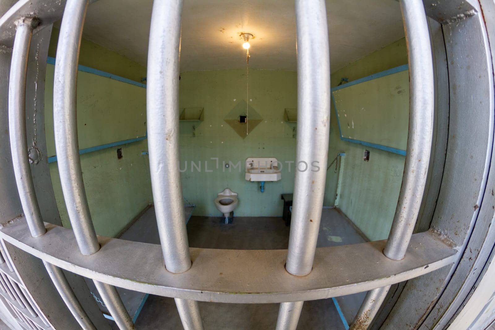 A jail cell with a toilet and sink. The cell is small and dirty. The bars on the window are rusty