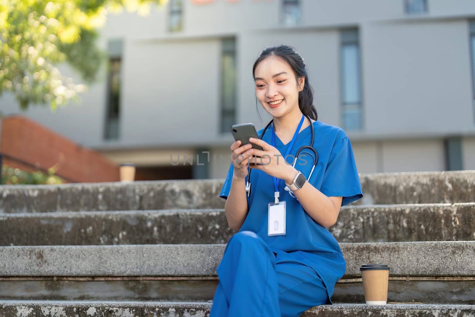 Cheerful Asian nurse in blue medical attire sitting on steps outdoors, using her phone during a break.