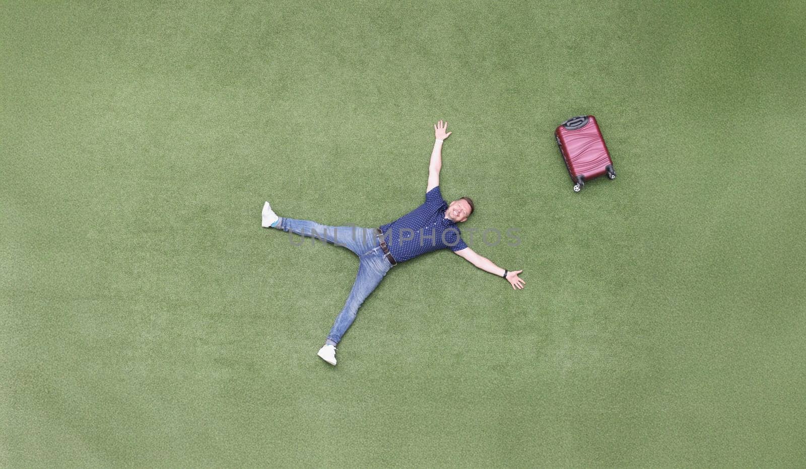 Happy man with a suitcase lies on a green lawn, top view. Arrival at the resort, vacation alone. Freedom from work