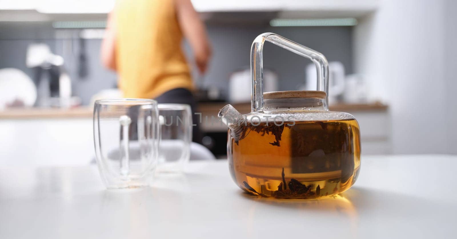 A woman made tea in a glass teapot, close-up, blurry. Glassware in the kitchen, coziness, hot herbal drink