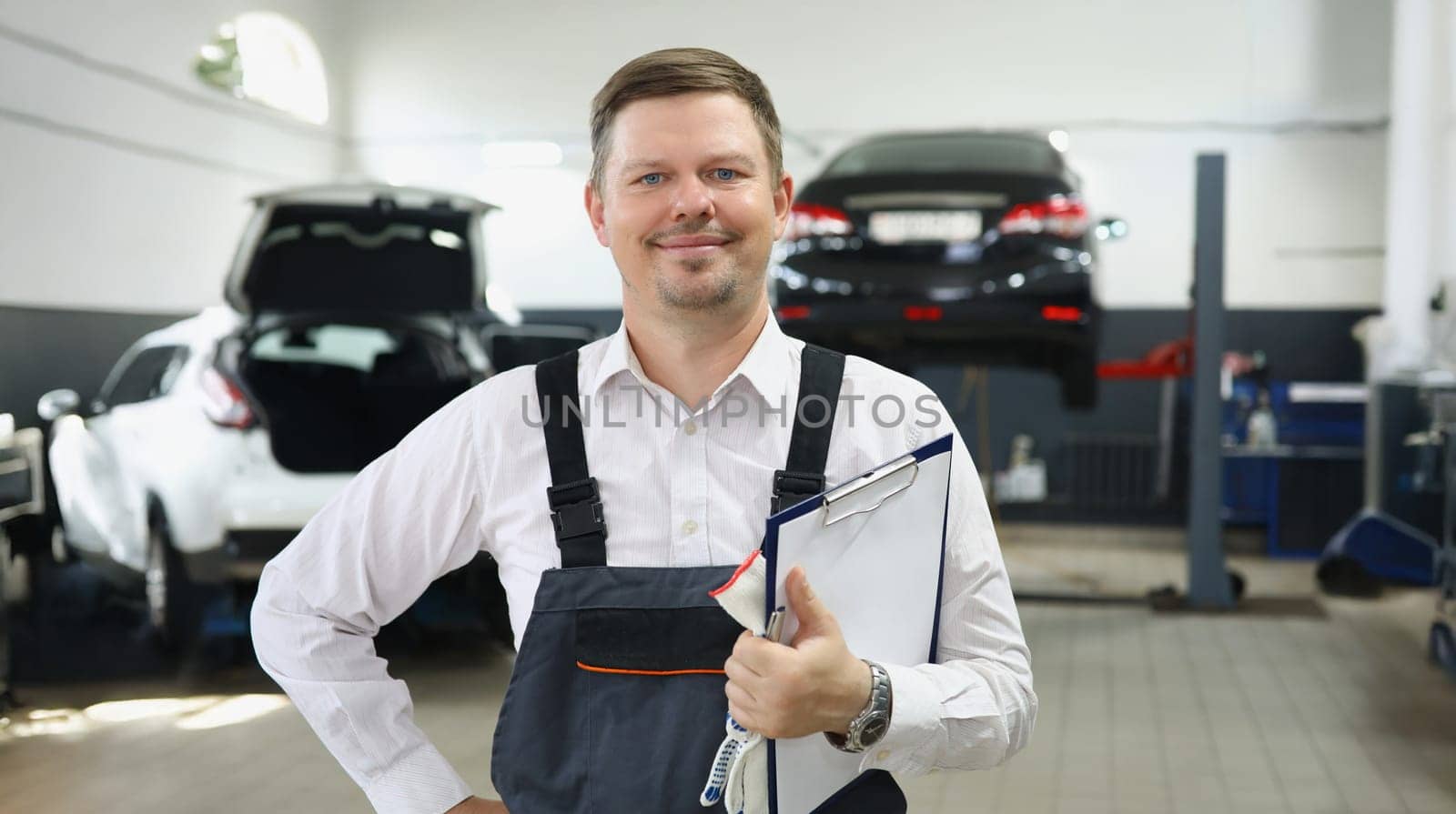 Portrait of qualified maintenance centre worker in special uniform posing on cars background. Mechanic male propose high service. Pit stop, garage concept