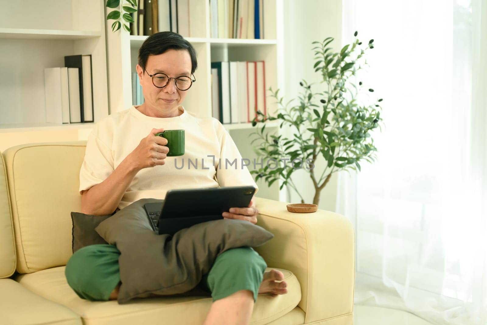 Happy middle aged man sipping coffee from a green mug and browsing on digital tablet.