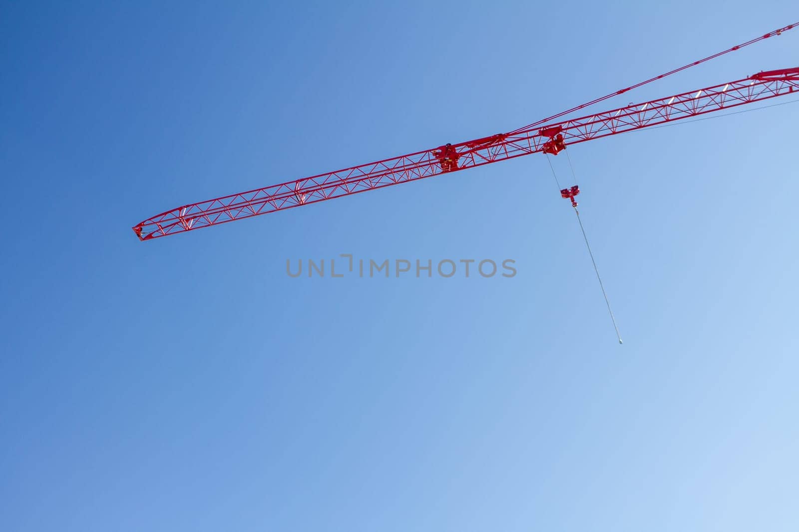 A crane is suspended in the air, with a red cable attached to it. The sky is clear and blue, with no clouds in sight. The crane is a large, red structure, and it is suspended high in the air