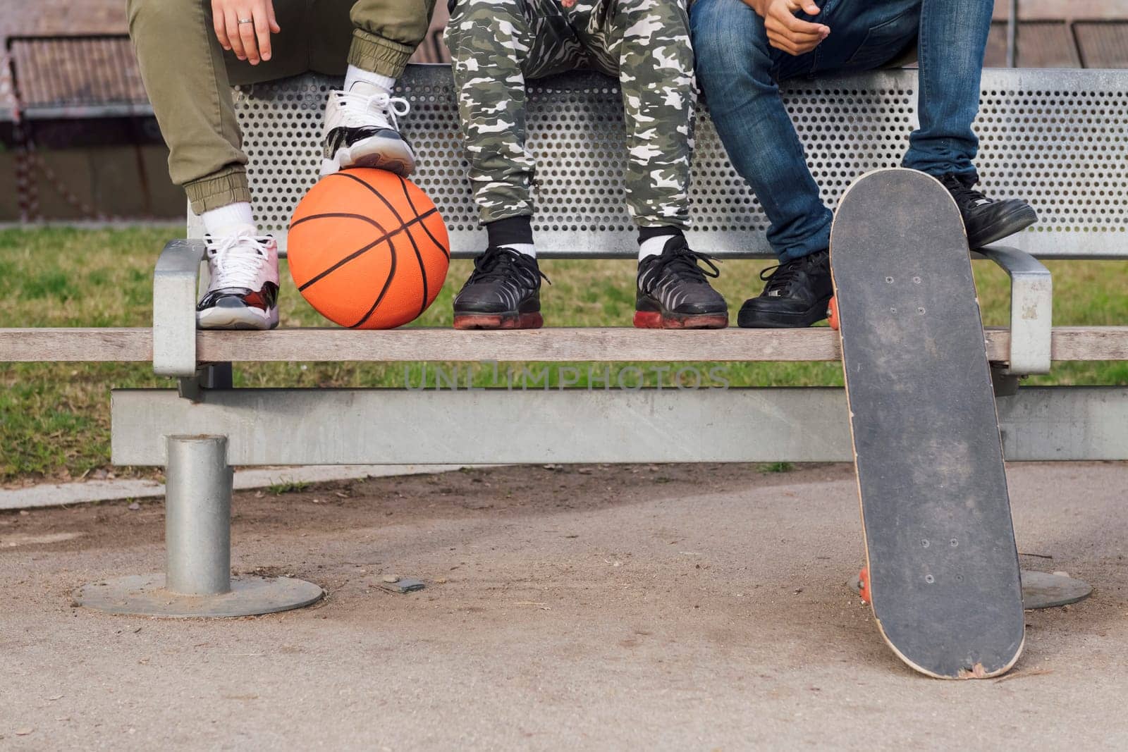 crop of three unrecognizable male friends sitting on bench of a park with basketball and skateboard, concept of youth and urban lifestyle