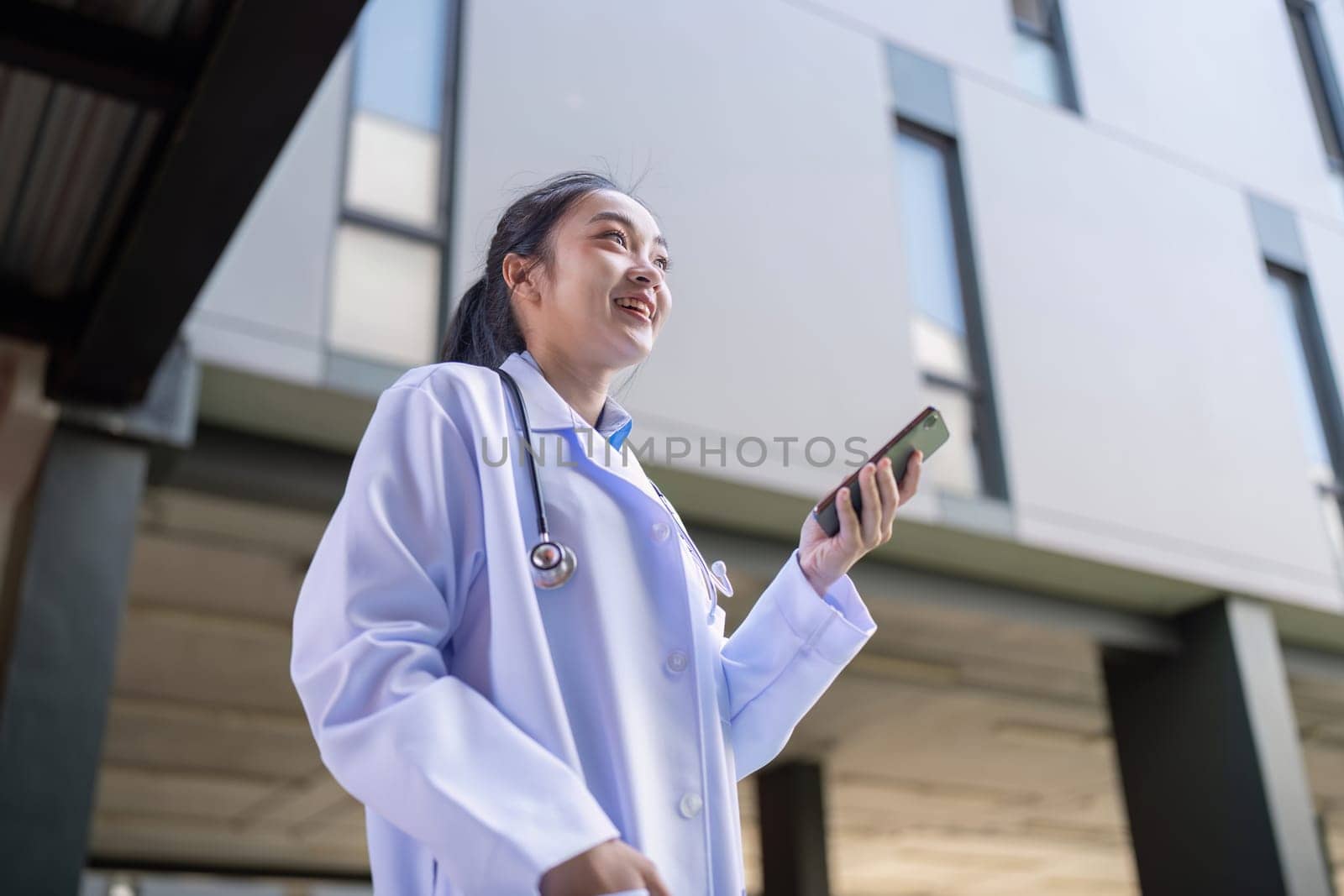 Asian female doctor wearing a lab coat, smiling while holding a smartphone outside, representing a blend of professionalism and communication.