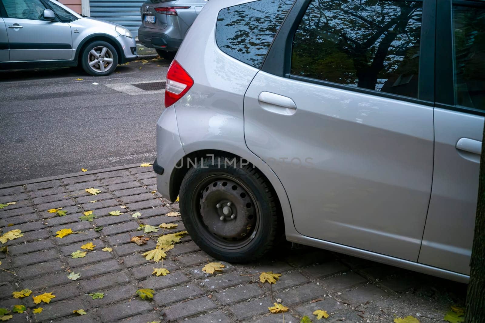 Lombardy, Italy - November 20th 2024 Parked silver honda jazz hybrid on a cobblestone street with fallen autumn leaves