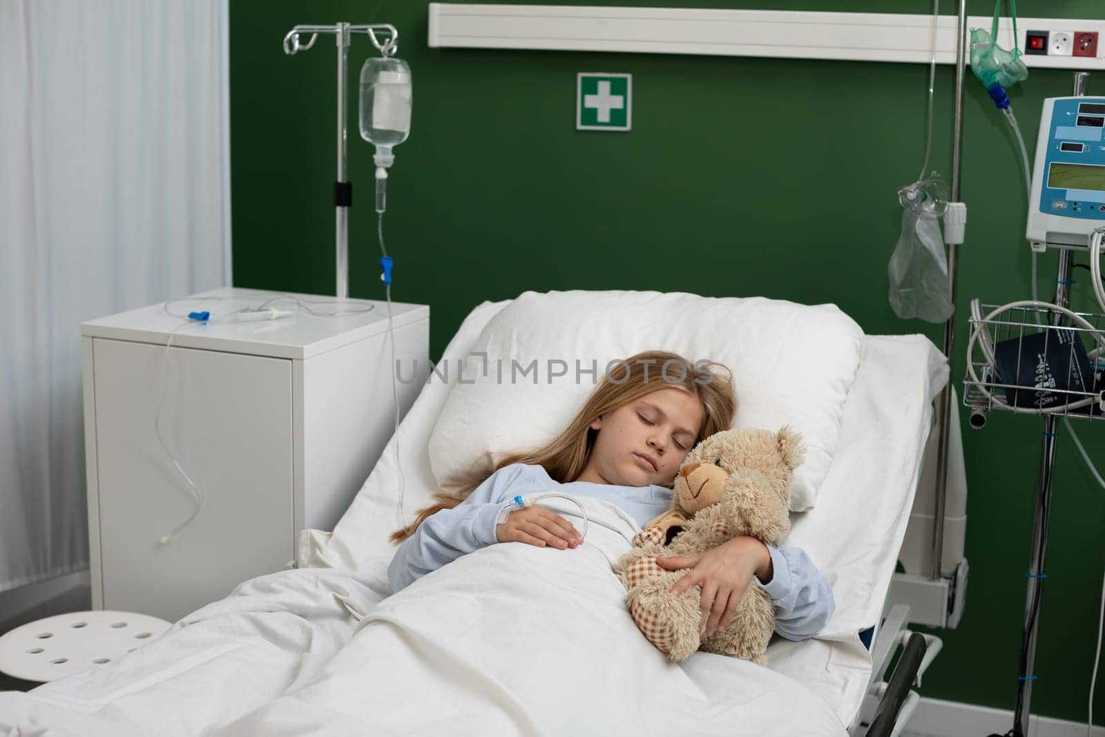 A girl rests in a cozy hospital bed, comforted by her teddy bear, showcasing love, care, and warmth