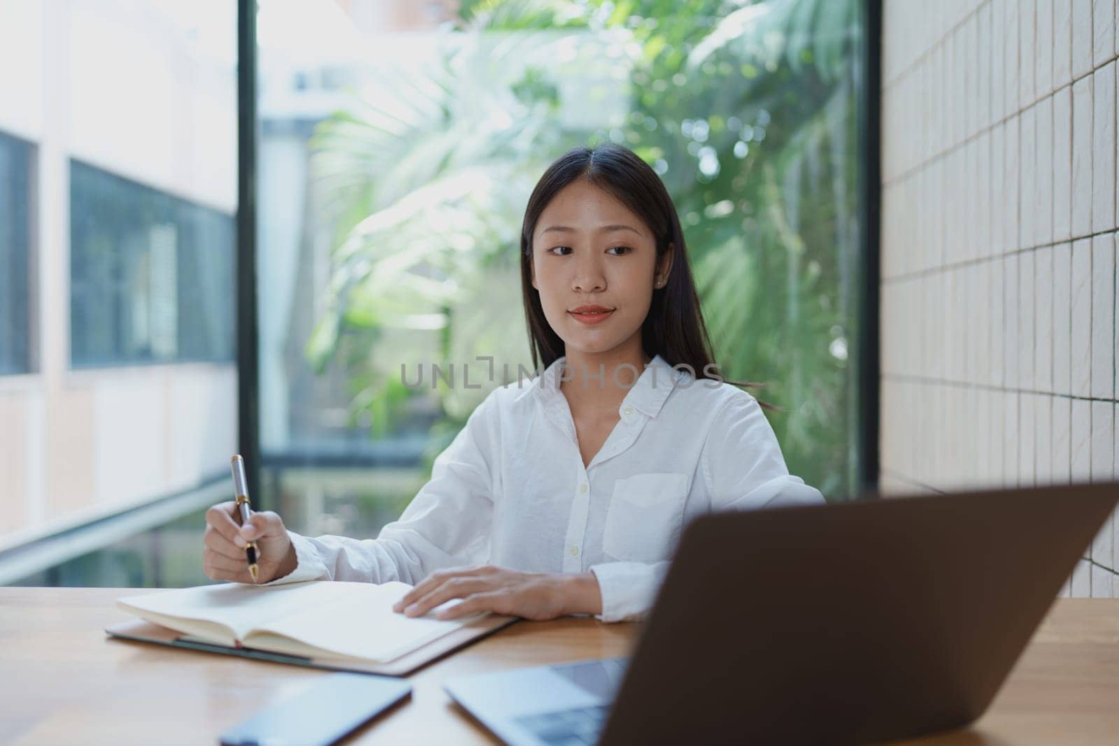 A young woman is seated at a wooden table in a bright and modern office, actively taking notes while using her laptop, representing focus and productivity in a contemporary workspace.