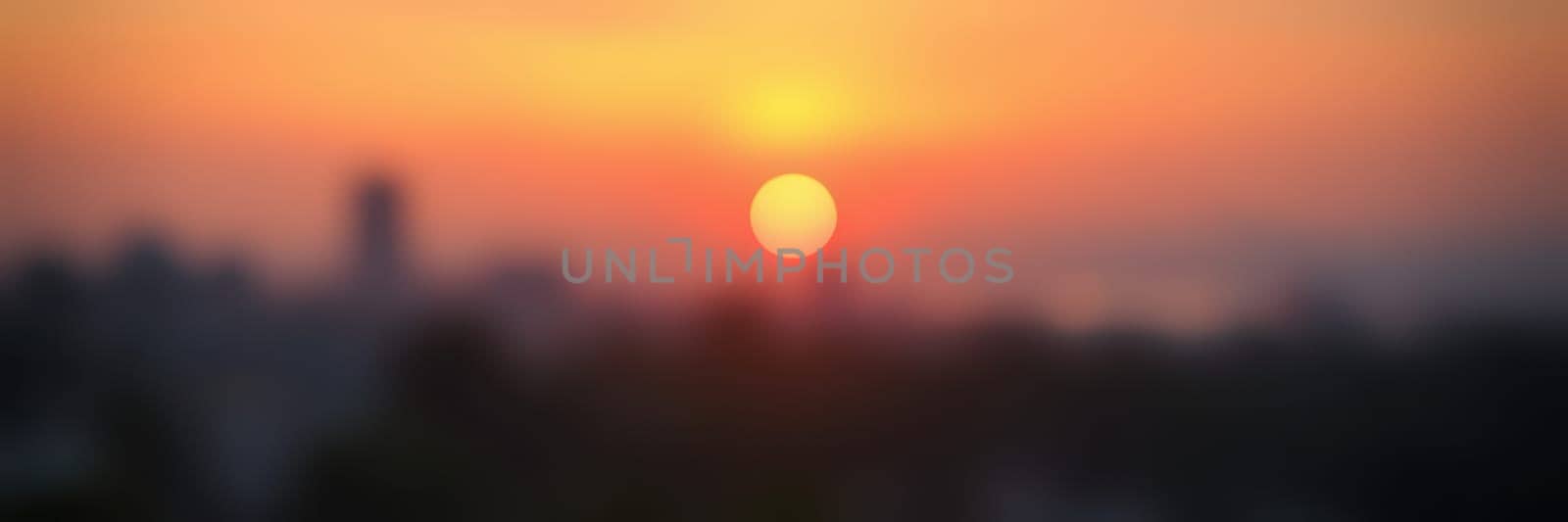 The sun sets behind the city skyline, casting warm orange and yellow tones across the sky. The soft glow illuminates the horizon, marking the end of the day.