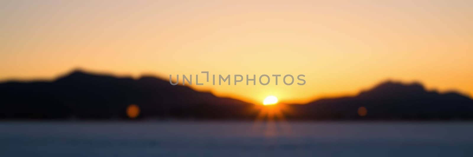 The sun rises slowly over mountain ranges, casting golden and orange hues across the calm lake's surface. Morning tranquility envelops the area as nature awakens.