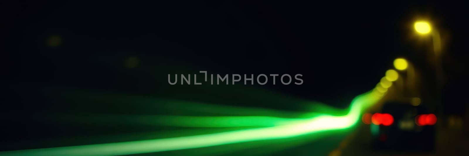 In the evening, a city street illuminates with warm streetlights while vibrant green light trails from speeding cars create a dynamic atmosphere.
