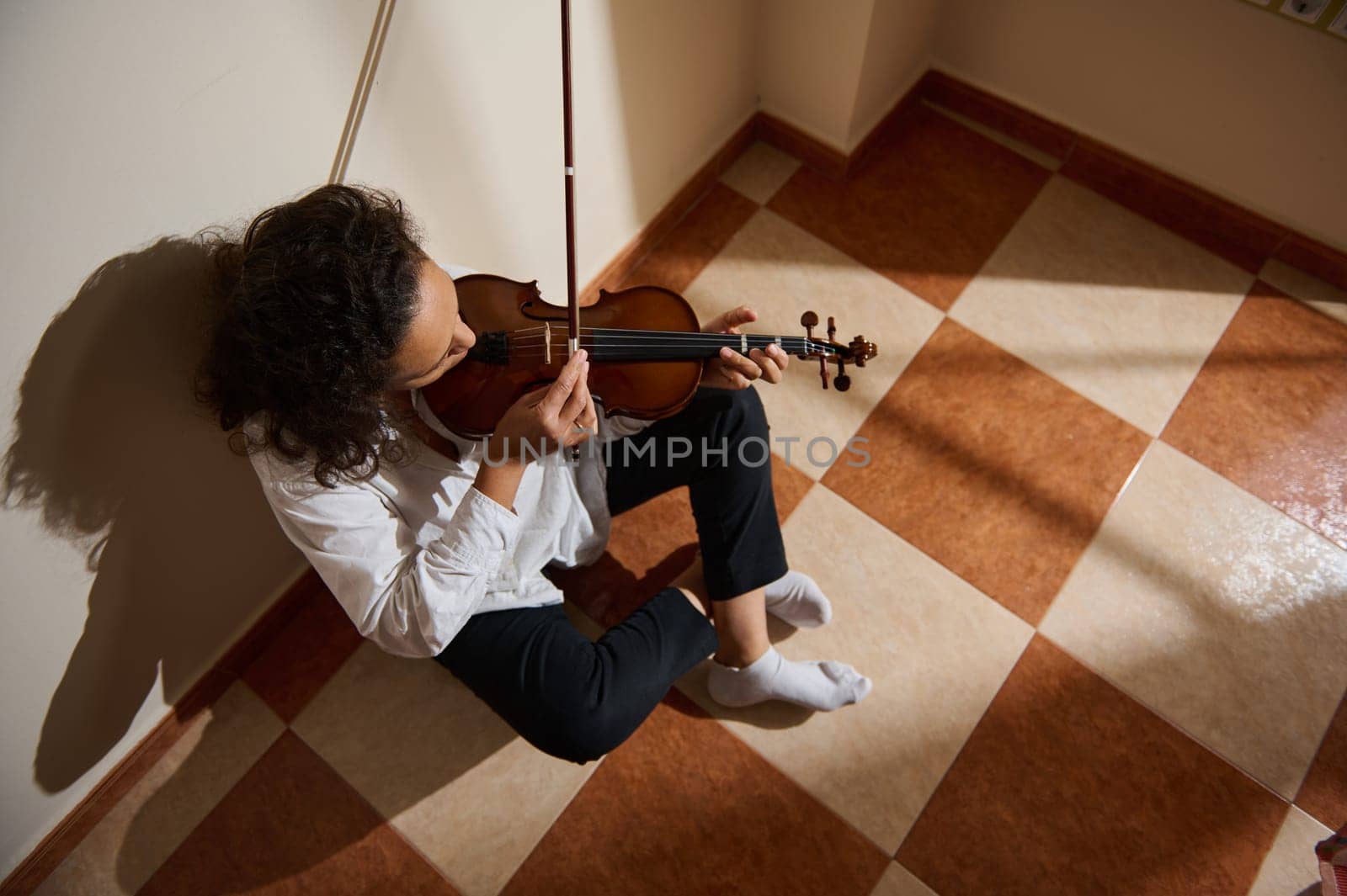 A musician seated on the floor plays the violin in a peaceful, sunlit room. The natural lighting enhances the tranquil atmosphere, creating a sense of calm and introspection.
