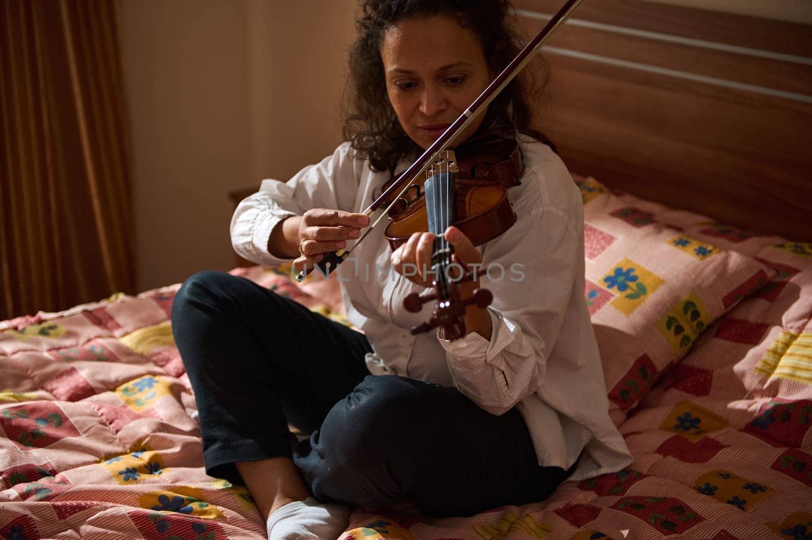 A woman in a casual white shirt plays the violin while sitting on a cozy bed, expressing concentration and passion for music.