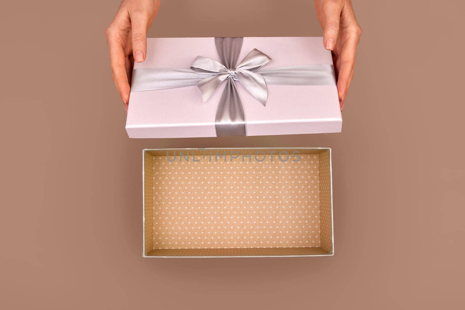 Female hands opening an empty gift box adorned with a silver ribbon bow set against a warm brown Mocha Mousse color of 2025 year background capturing the essence of surprise and celebration from above