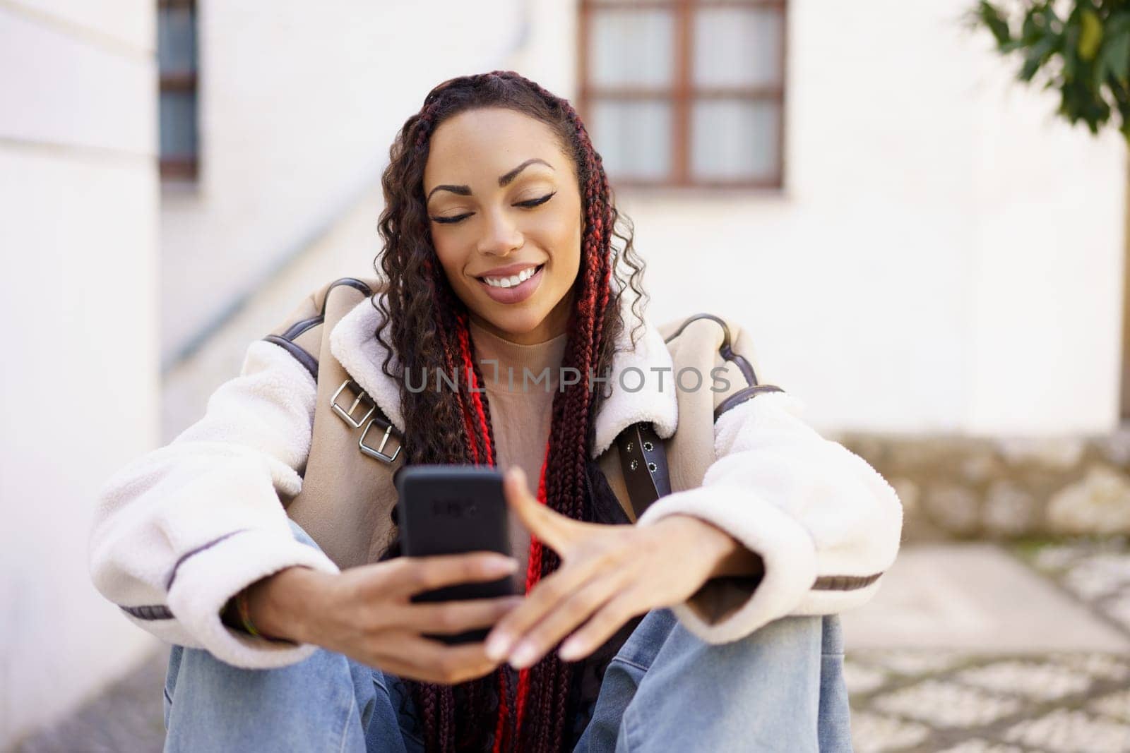 In a lively outdoor setting filled with sunshine, a cheerful young woman with beautifully braided hair radiates happiness as she happily smiles while using her smartphone, enjoying a joyful moment