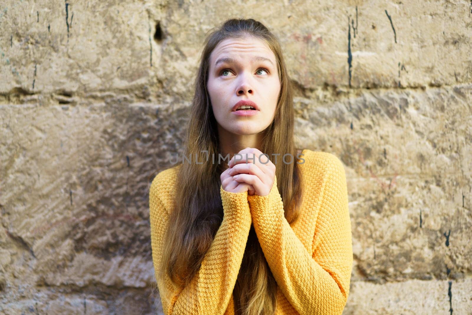 A young woman in a vibrant yellow sweater gazes upward with hope, her emotions beautifully reflected. The rustic stone wall adds depth, creating a striking visual of peace and inspiration