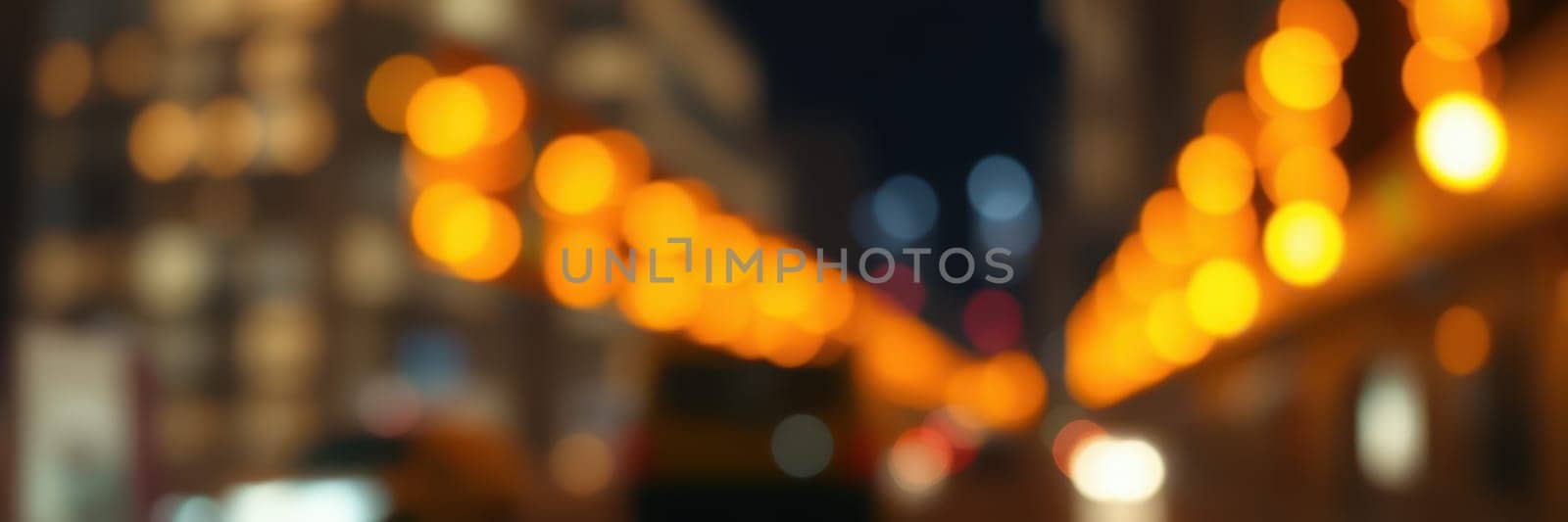 Softly blurred lights illuminate a city street at night, creating a warm and inviting atmosphere. Buildings line the street, enhancing the urban setting and evening ambiance.