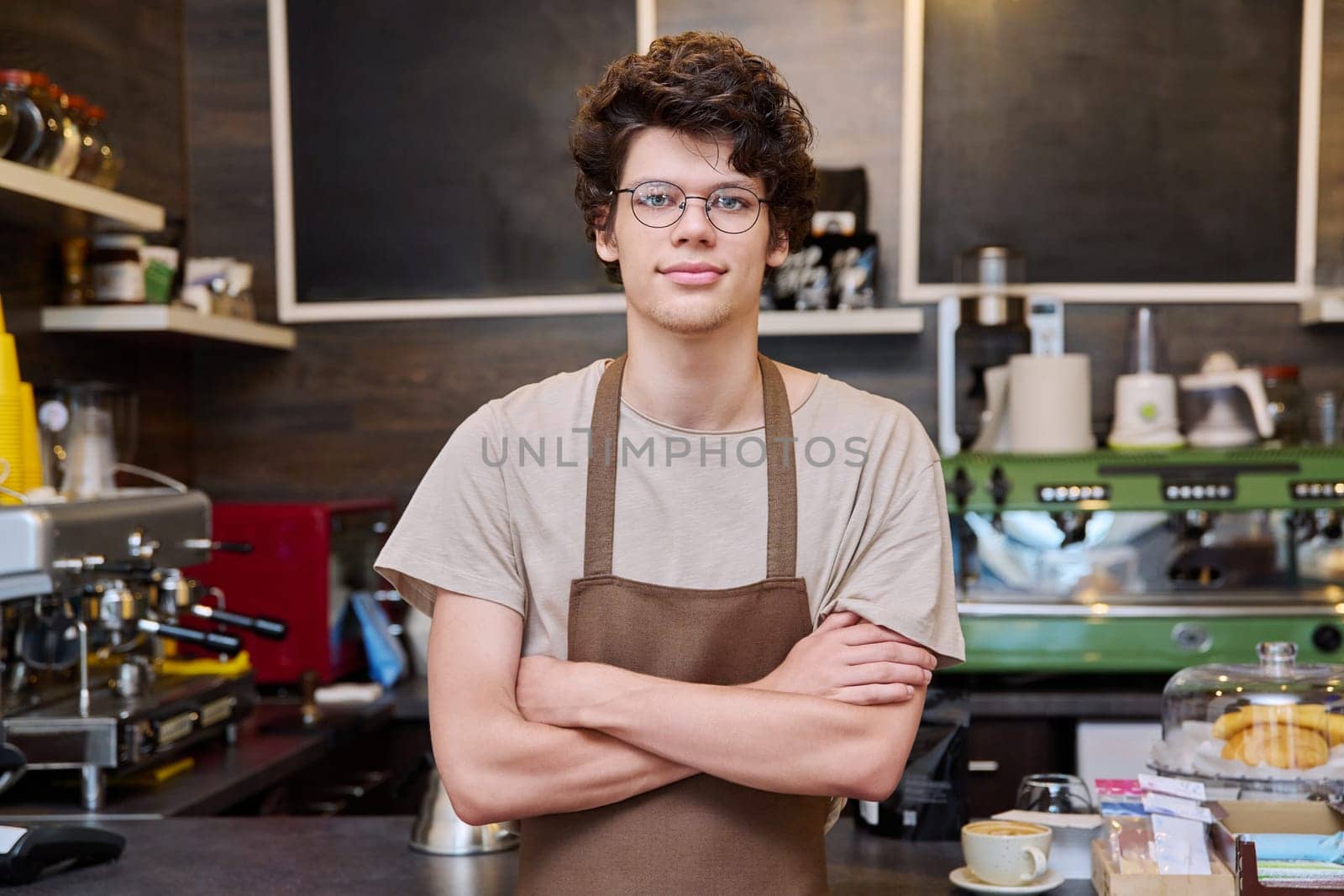 Young confident male coffee shop worker with crossed arms by VH-studio