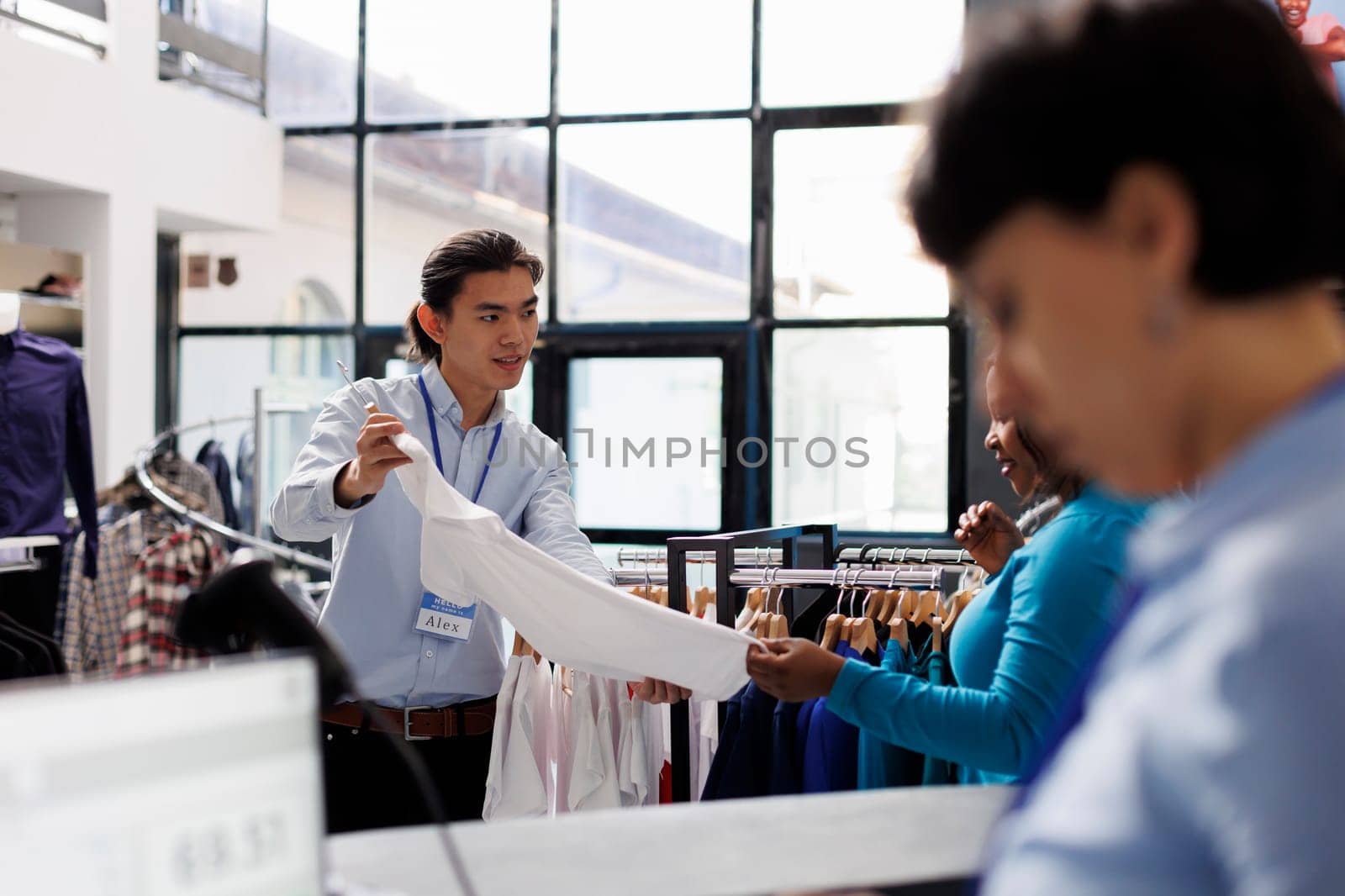 Asian employee helping woman shopper with elegant clothes, discussing clothes material in modern boutique. African american customer shopping for casual wear, looking at stylish merchandise