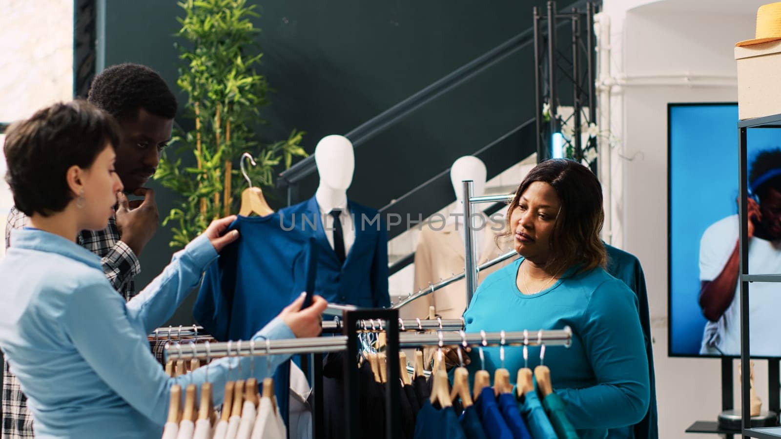 Caucasian worker helping cheerful couple with stylish merchandise, discussing items material in clothing store. African american customers shopping for fashionable clothes in modern boutique