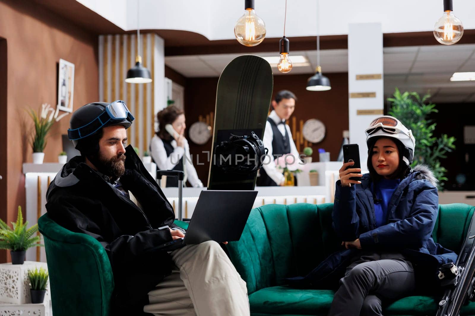 Boyfriend using laptop and girlfriend with mobile phone enjoying digital communication. Wintersports enthusiasts in hotel lobby for ski and snowboarding fun waiting with their smart devices.